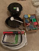 Black speckled enamel canner with lid, oval roasting pan with rack, white plastic ladle, disposable aluminum pans stacked, box with various canning and kitchen supplies including glass jars and lids, and packaged BBQ tools on carpeted floor.