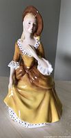 Front view of Royal Doulton Sandra porcelain figurine showing seated woman wearing yellow dress and brown hat.