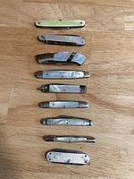 Top-down view of 9 abalone folding knives arranged vertically on wood surface showing their handles and size variety.