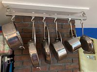 Six All-Clad cookware pieces hanging on rack against brick wall