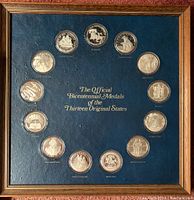 Overall front view of oak frame holding all 13 official bicentennial state medals in protective cases around central gold-foil title on blue felt backing.