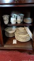Shelved china showing stacked plates, teacups, and saucers with holly pattern and gold trim.