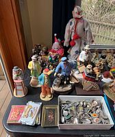 Wide view of a collection of clown figurines including Emmett Kelly clowns, assorted souvenir spoons in box, and playing card decks on a table.