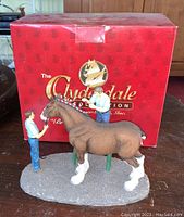 Figurine with two men braiding a brown Clydesdale horse in front of red branded box