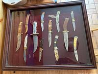 Full view of all 12 knives arranged in wood-framed glass case with burgundy background.