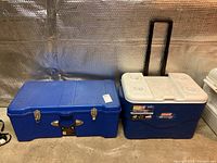 Blue Contico plastic storage locker with metal closures and handle next to blue Coleman Extreme5 cooler with white lid and telescoping handle