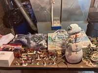 Wide shot of assorted holiday village pieces on wooden table, including miniature figurines, trees, large snowman, boxes, and electrical cords.