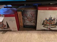 Three boxed Department 56 items side by side on a shelf: red/white Discover series box, North Pole Woods Oakwood Post Office Branch box, Sleigh Launch building box