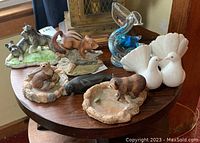 Overview of animal figurines on round wooden table including otter, dogs, chipmunk, seal, raccoon on dishes, two doves and a blue glass pelican figurine