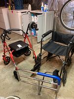 Photo showing red walker with padded seat, black mobility chair, metal cane, and metal bed rail in basement setting.