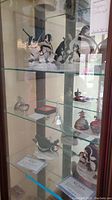 Glass display cabinet with multiple penguin figurines on upper shelf and pewter Budweiser eggs and bells on lower shelves.