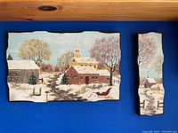 Two paintings on wood panels depicting winter village scenes with snow, trees, and buildings, set on a wooden surface.