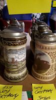 Photo showing front view of two West German steins with pewter lids, one with Monaco cityscape and one with locomotive image, on shelf with sticky notes labeled 'West Germany'
