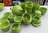 Full set of Fiesta ware items in chartreuse green including teapot, mugs, gravy boat, cream and sugar with resting dish, and teacups, arranged on a white table.