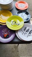 Top view of decorated Halloween Fiesta Ware plates including orange with black cat, black with ghostly cat, green with 'BOO' and black cat, purple with bubbling cauldron, and white with dancing skeletons.
