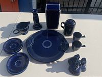 Set of various cobalt blue Fiesta ware pottery pieces displayed on white table under sunlight showing platter, saucers, creamer, shaker, chocolate pots, candle holders, napkin dispenser