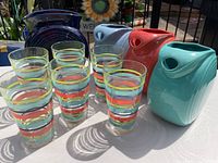 Set of 6 banded color Fiesta Ware glasses with 3 disc pitchers and cobalt dish vase on table outdoors.