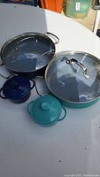 Large cobalt blue and turquoise cast iron cookware with glass lids and small matching mini cocottes on white table surface.