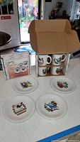 Photo showing four plates and four mugs from Fiesta Twelve Days of Christmas set, numbered 1 to 4, displayed on a kitchen counter with some items still boxed.