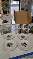 Photo showing four salad plates laid out on a surface with numbers 9 through 12 and matching mugs in original packaging stacked next to plates.