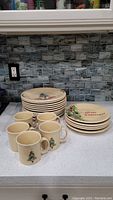 Grouped photo showing 5 cream-colored ceramic mugs with Christmas tree motifs and approximately 15 matching plates stacked separately, all on a white countertop.