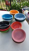 19 Fiesta Ware ceramic bowls in various colors arranged on a white round table outdoors, visible colors include blue, orange, red, green, black, pink, yellow, white, turquoise.