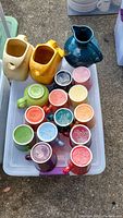 Top view of Fiesta Ware ceramic mugs and pitchers arranged in a plastic bin showing various colors and styles.