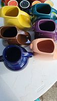 Top and angled views of five disc-shaped Fiesta Ware pitchers in vibrant colors: yellow, brown, pink, teal, purple.