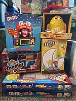 Photo showing six M&M collectible boxes including the blue animated clock radio box with Red M&M figure, yellow character bowl box with seated Yellow M&M, goose neck desk lamp box, candy dispenser truck box, and two Super Draw drawing board boxes.