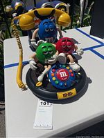 Front and side view of M&M's animated telephone featuring Red, Green, and Blue M&M character figures on the base, yellow handset connected by coiled yellow wire, and colorful push-button rotary dial with 'REDIAL' and 'FLASH' buttons.