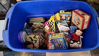 Blue tote filled with various vintage metal lunchboxes, tins, and plush toys featuring characters from Peanuts, Rugrats, and Looney Tunes.