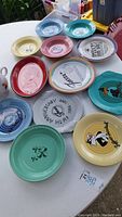 Various Fiesta brand plates and platters on white table including Warner Bros cartoon characters, anniversary plate, and colorful Fiesta designs