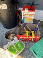 Photo showing garden tools including pruners, shears, hand tools, wooden clothespins in a metal container, a green garden kneeler, and a box previously holding round containers.