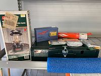 Photo showing Coleman gas lantern boxed, green Coleman 425 stove with latch closed, solar powered radio, several foil pans, and other camping utensils placed on metal shelf