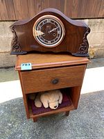 Front view showing the ForestVille mantle clock placed on the wooden side table with a drawer, plush dog toy, and book visible in the open shelf below.