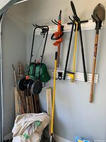 Wide view of the garden tools hung on wall hooks, showing multiple garden tools including a green spreader, shovel, rake, bamboo sticks, trimmer and other tools.