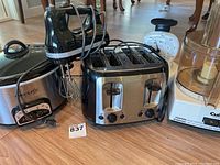 Full view of all four appliances on floor showing Kitchenaid hand beater, Crockpot slow cooker, Black & Decker 4-slice toaster, and Cuisinart food processor container.