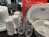 Photo showing stacked white Corelle plates and bowls, white mugs, clear faceted tall glass drinking glasses, CorningWare casserole dish, and Betty Crocker's Cookbook with red cover in background.