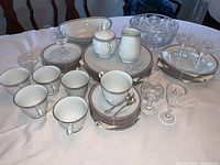Full display of the dinnerware and glassware set showing plates stacked in groups, teacups arranged in front, the cream pitcher and sugar bowl placed on plates, an oval serving dish and a crystal bowl with 6 crystal glasses around.