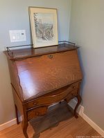 Front angled view of vintage wooden secretary desk showing slant-front, drawers, and ornate edges.