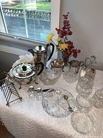 View of antique silver plated tea and coffee set with black handles, crystal bowls, lidded jars, and salt and pepper shakers on a table with white cloth and decorative flowers in background