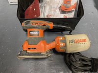 Ridgid square power sander with dust collection bag next to black soft carrying case and instruction manual inside case.