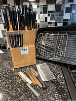 Front view of wooden block filled with black-handled knives, kitchen shears, several loose knives, and roasting pan with racks visible behind.