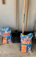 Three snow shovels with wooden handles standing upright next to two full bags of blue and orange packaged Alaskan Pet Friendly Ice Melt in garage.
