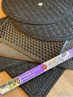 Multiple black non-slip rubber backed door mats stacked showing different carpet textures and honeycomb patterned mat with holes; Gila Privacy Control window film box in foreground.