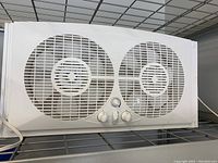 Front view of a white twin window fan with two large circular fans and three control knobs on a metal shelf.