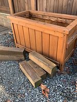 Wooden planter with four wooden blocks in front on gravel ground near wood fence