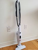 Front view of the white 3-in-1 vacuum cleaner with cord wrapped around handle resting on wooden floor near wall.