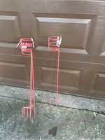 Two red beverage holder ground stakes leaning against a garage door on pavement.