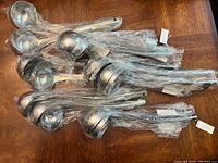 Sixteen large stainless steel ladles wrapped in plastic laid out on a wooden surface, showing handles and bowls piled together.